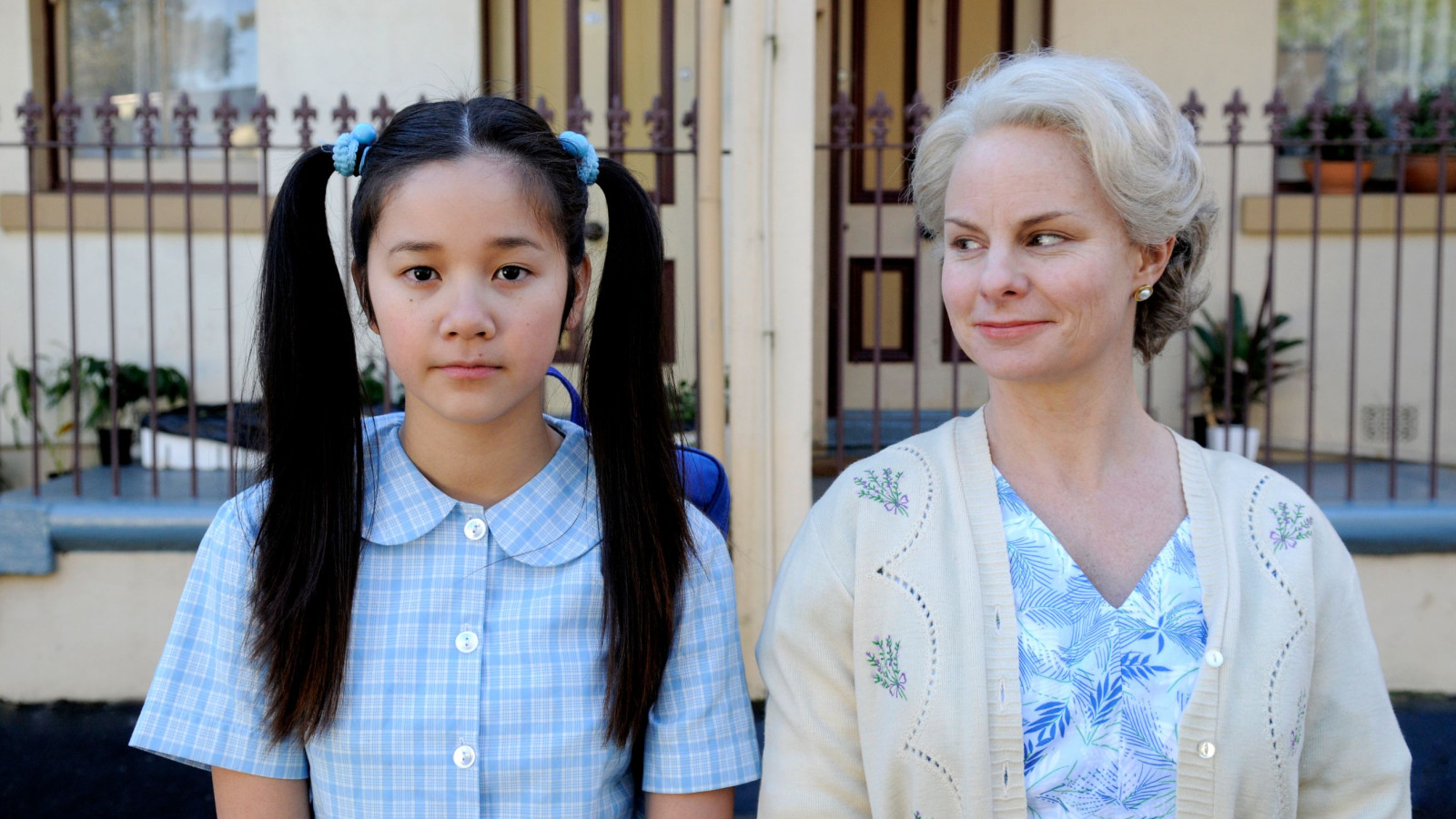 A young girl with long, dark hair tied into pigtails wears a blue checkered dress and looks solemn, standing next to an older woman with short, light hair and a warm smile, dressed in a floral blouse and cardigan. The background features a house with two doors and a decorative fence, hinting at a suburban setting.