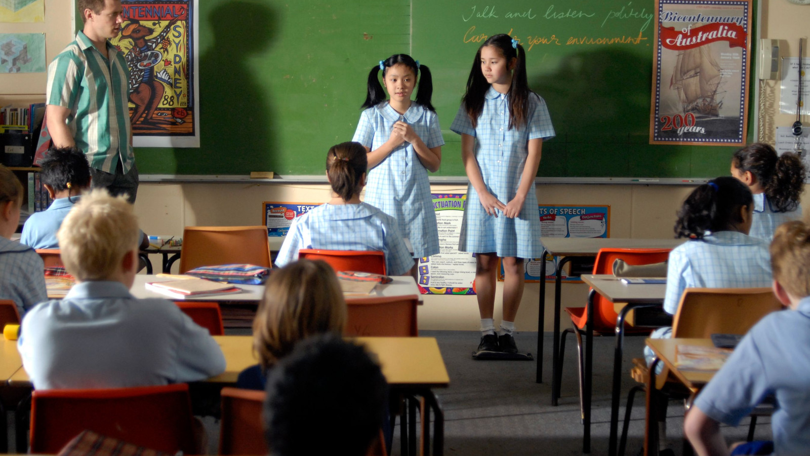 A classroom scene features a teacher and two students in blue uniforms standing in front of a blackboard, engaging in a presentation. The seated students face them, appearing attentive, as educational posters decorate the walls, emphasizing a school setting.