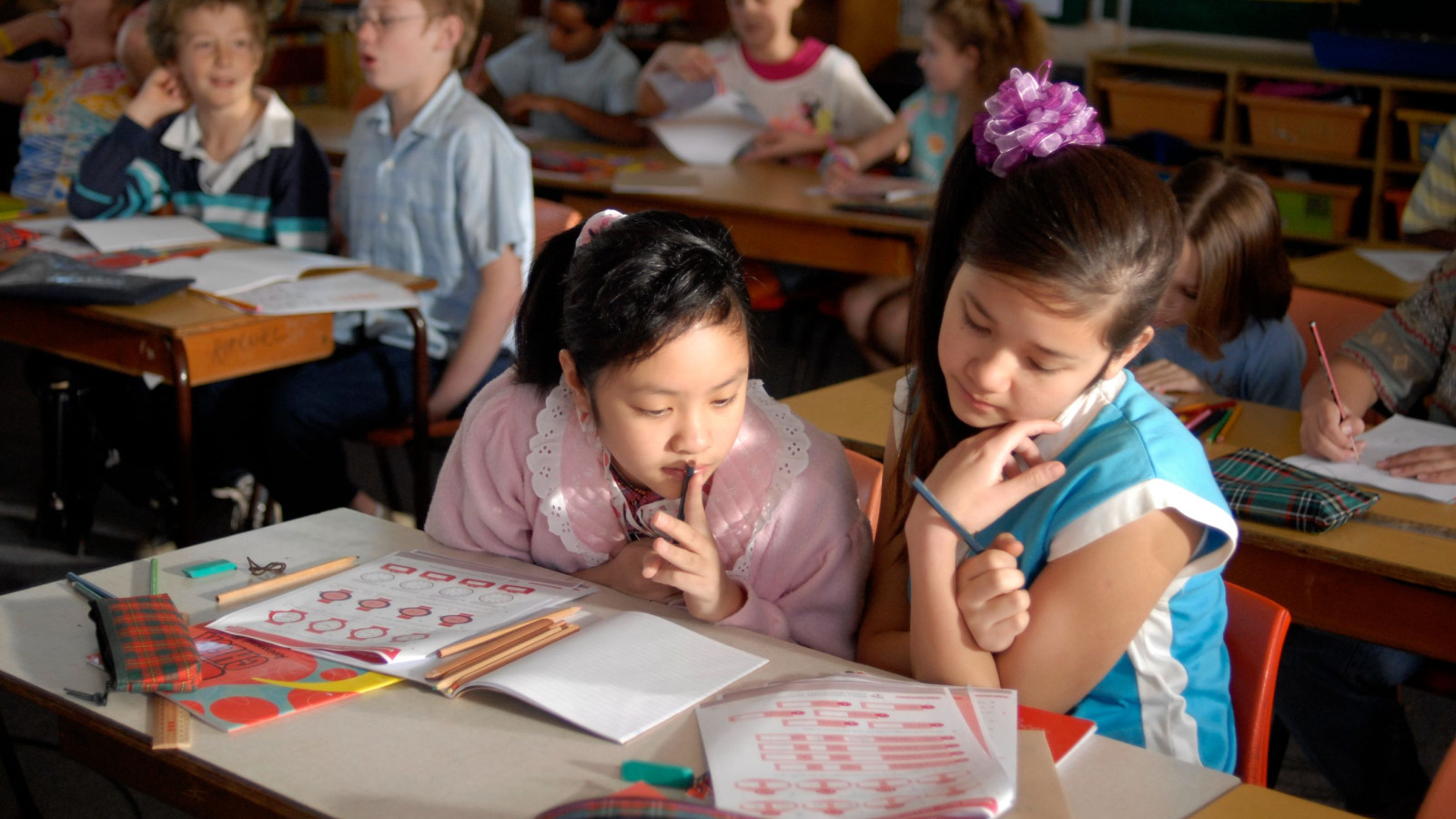 Two girls sit at a desk in a classroom, focused on their work. One girl wears a pink sweater with a lace collar and holds a pencil to her lips, while the other, dressed in a blue top, leans in to examine their papers, which feature various shapes and colors. In the background, several other students are engaged in their activities.