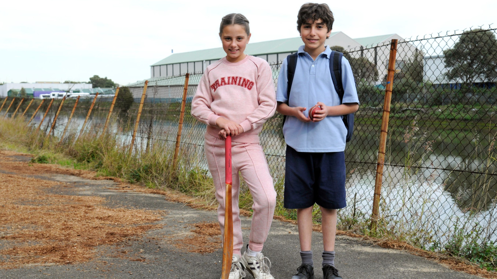 Two children stand on a path by a fenced area near a body of water. A girl in a pink tracksuit holds a cricket bat, while a boy in a blue shirt and shorts holds a cricket ball and has a backpack on.