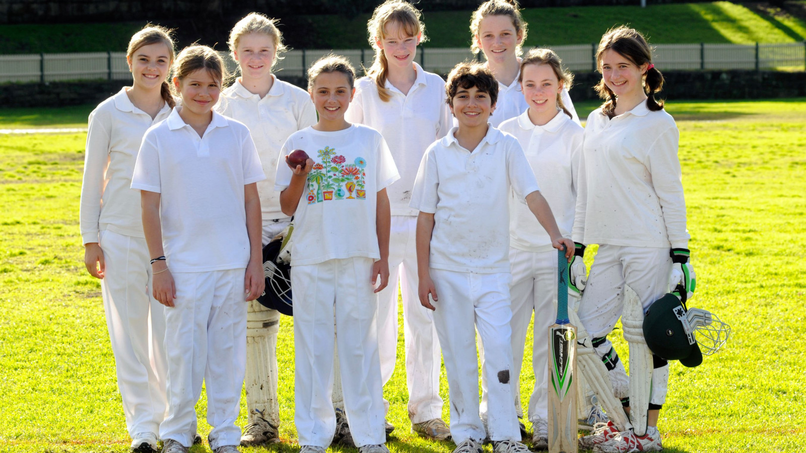 A group of ten young cricketers stands on a grassy field, wearing white cricket uniforms. A boy holds a cricket ball, while another girl displays a bat and gloves, with smiles on their faces as they pose together in bright sunlight.