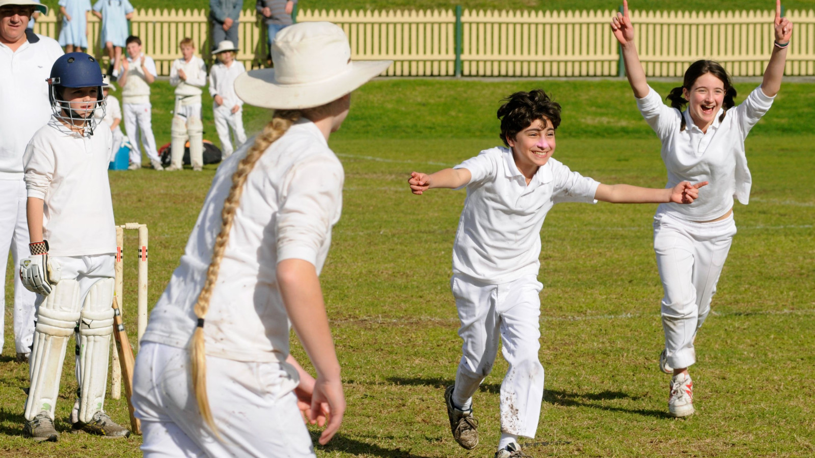 A group of children in white cricket uniforms celebrate joyfully in a grassy field, with two kids running towards the camera, arms raised in excitement. In the background, onlookers and players watch the celebration, highlighting a vibrant atmosphere of sport and camaraderie.