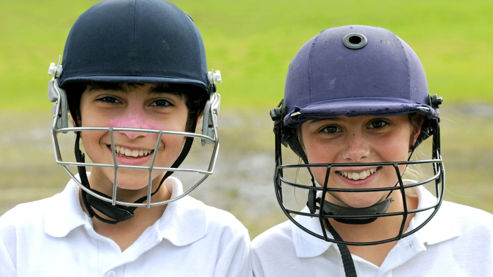 Two smiling young players wearing cricket helmets stand side by side, showcasing their enthusiasm for the sport. They are dressed in white shirts, and the background features a lush green field.