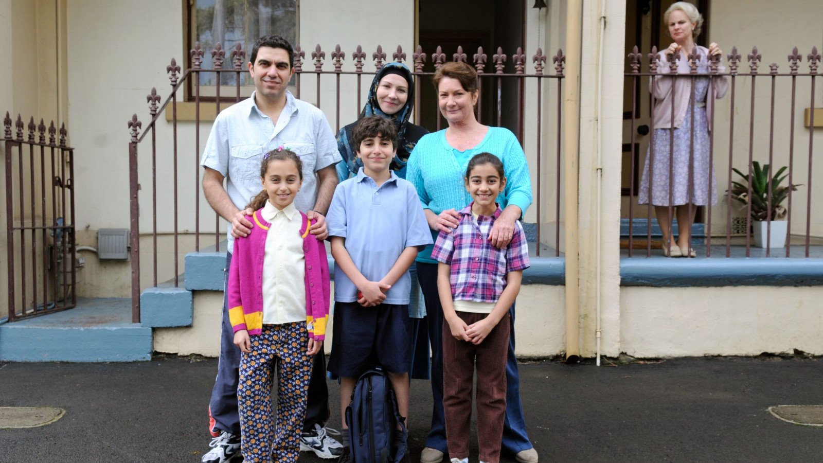 A diverse group of six people poses together in front of a house. The scene features three adults, two girls, and a boy, with the young children dressed in colorful clothing. The adults appear to be smiling and relaxed, while a woman stands in the background, looking on from the porch. The setting includes a well-kept yard and decorative plants.