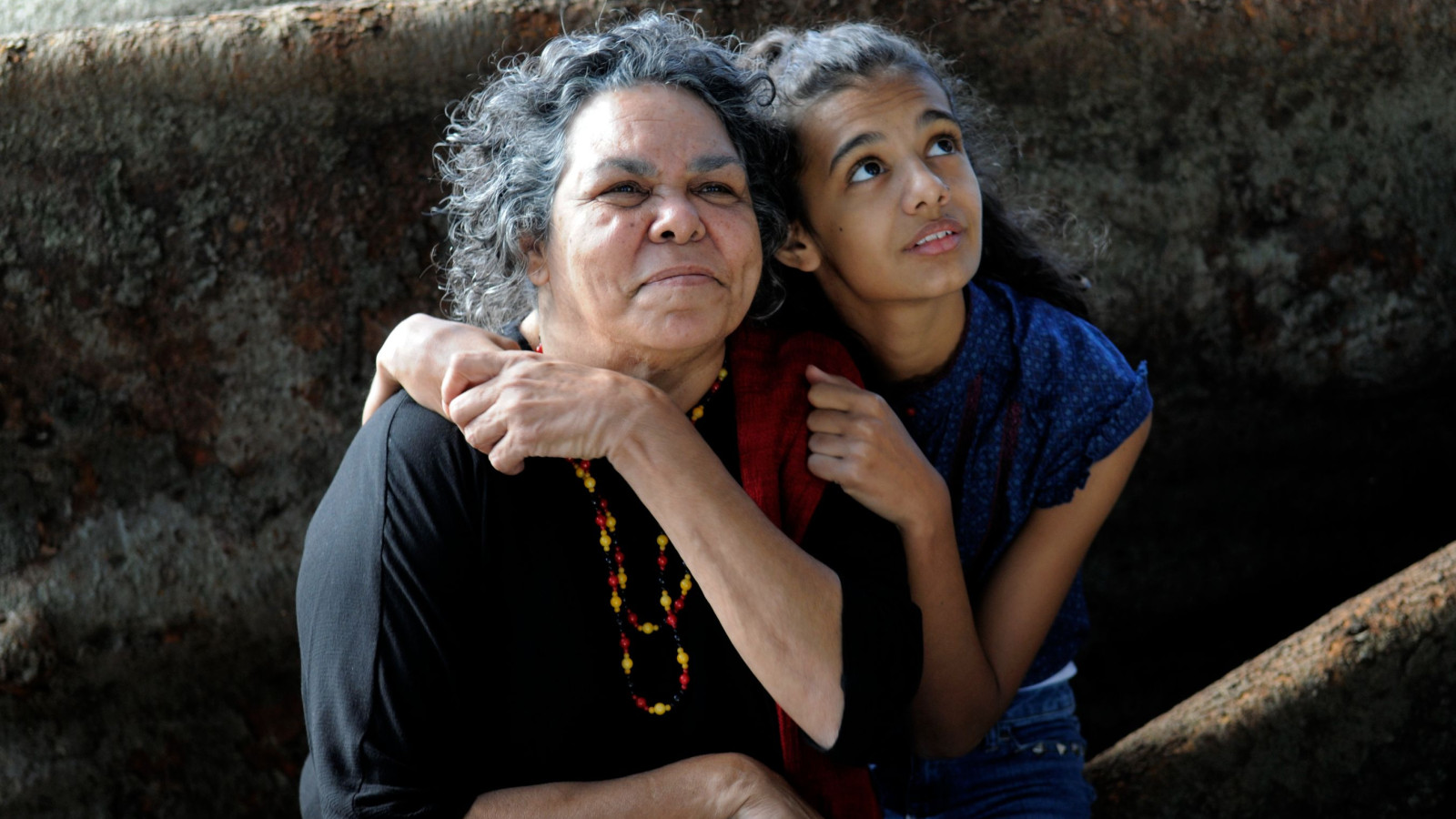 An elderly woman and a young girl share a moment of affection, with the girl resting her head on the woman's shoulder while looking upward with a thoughtful expression. The woman smiles gently, wearing a necklace of colorful beads, against a rustic backdrop.