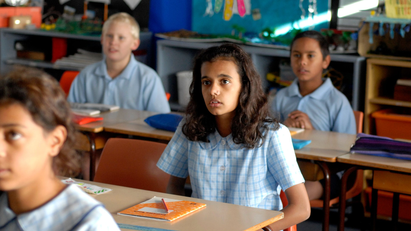 A classroom scene features several children seated at desks, wearing light blue school uniforms. The focus is on a girl with curly hair, looking attentively ahead, while other students are engaged in their seats. The background shows colorful educational materials on the walls.
