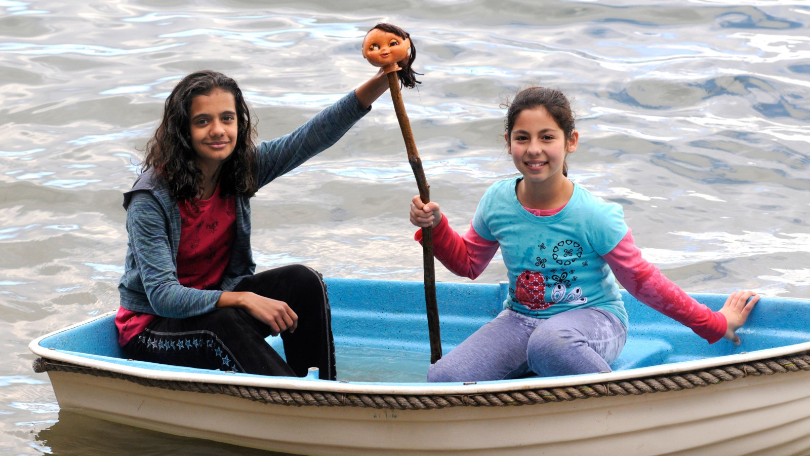 Two girls are sitting in a small boat on a body of water, smiling at the camera. One girl is holding a stick with a doll's head attached to it, while the other is sitting casually with her hand on the boat's edge. The water reflects the surroundings softly.