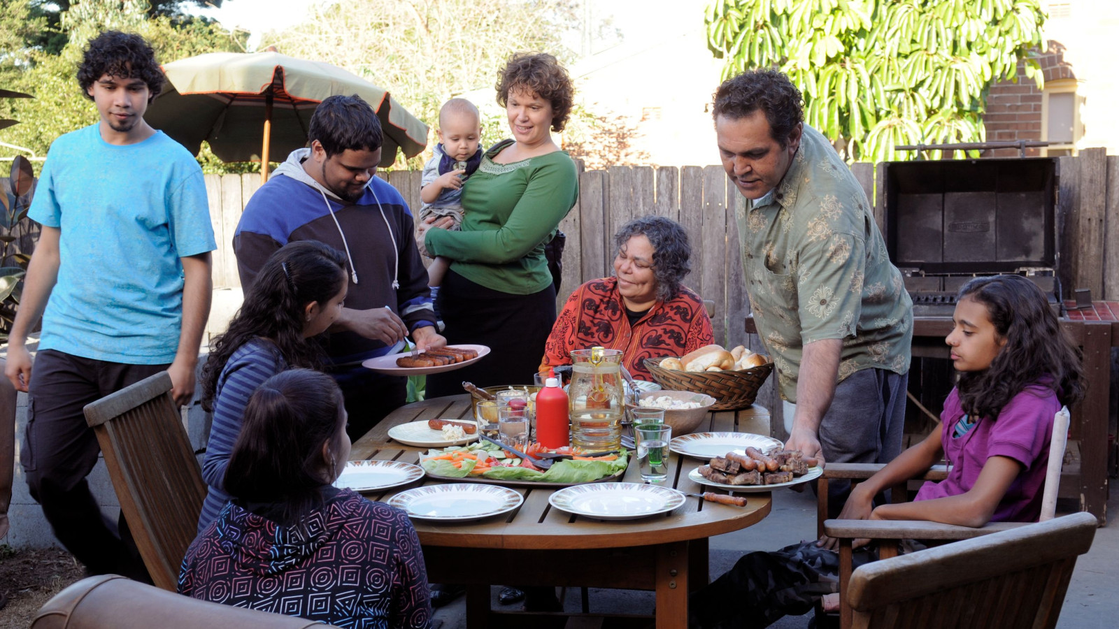 A diverse group of people gathers around a wooden table set with various dishes, including grilled meats and salads, in a backyard. Someone is serving food, while others chat and hold a baby, enjoying the outdoor setting surrounded by greenery and a grill in the background.