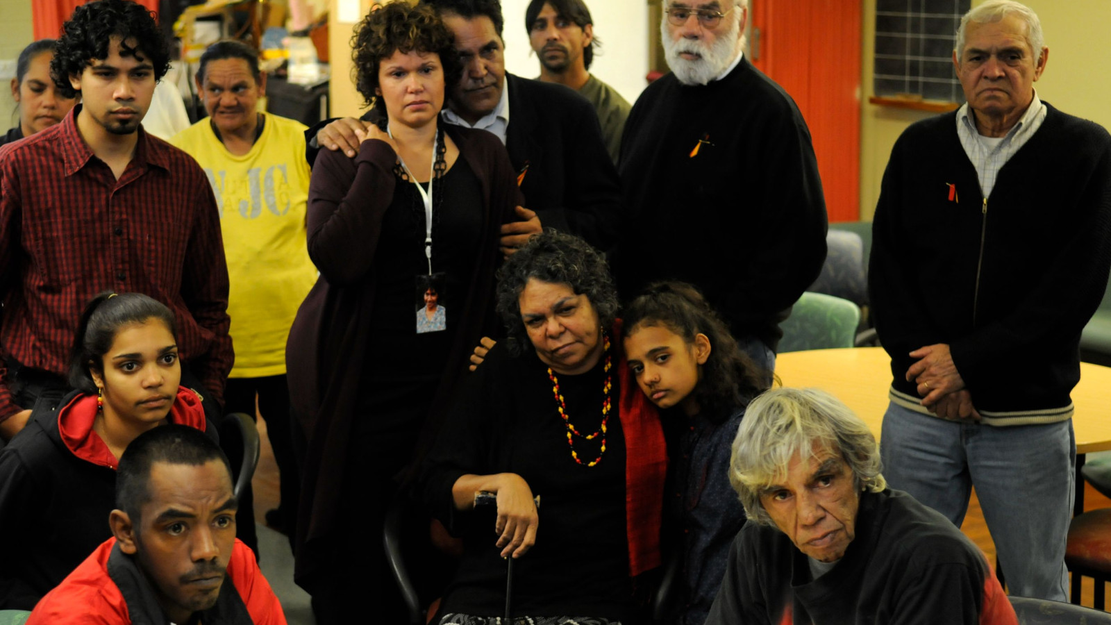 A diverse group of people stands and sits together in a communal space, showing expressions of concern and reflection. Some individuals are wearing colorful clothing, while others have more subdued attire, and there are items like a table and chairs in the background, suggesting a gathering or meeting environment.