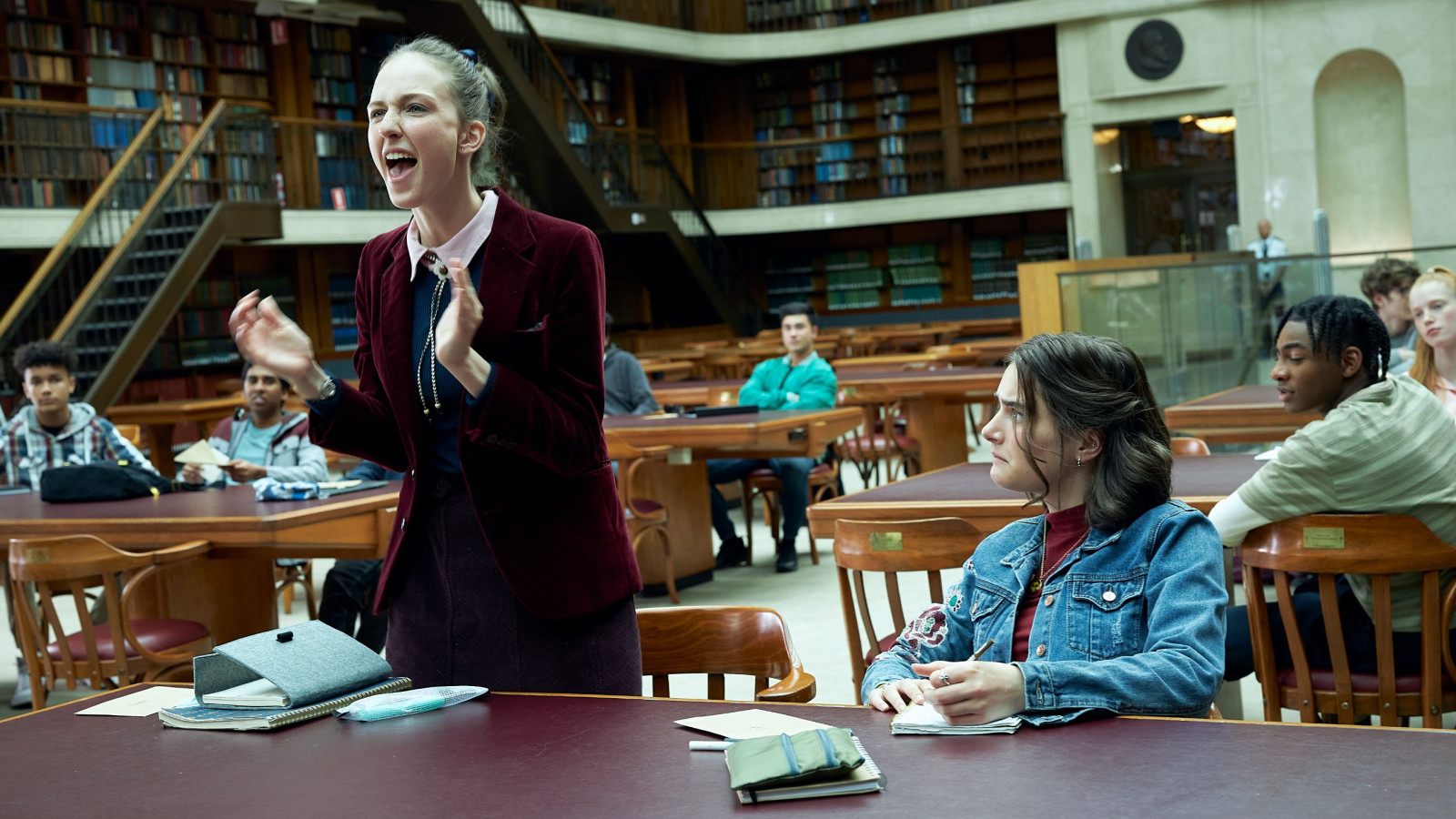 THE PM'S DAUGHTER_Sadie (Natalie English)_Cat (Cassandra Helmot)_Ep3_SD33_0105 A woman in a burgundy blazer passionately addresses a group of mostly young people seated at tables in a library, while they observe her attentively. The setting features wooden tables and shelves filled with books in the background.