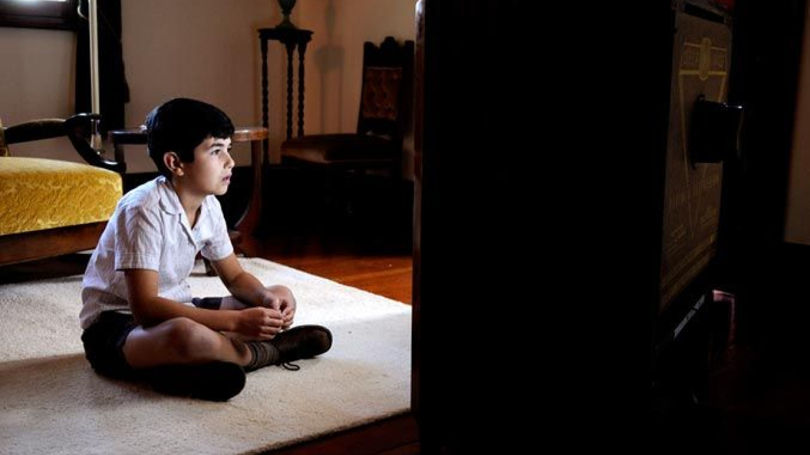 A young boy sits cross-legged on a rug in a dimly lit room, intently focused on an old television set in front of him. Behind him, a vintage chair and a table add to the nostalgic atmosphere of the space.