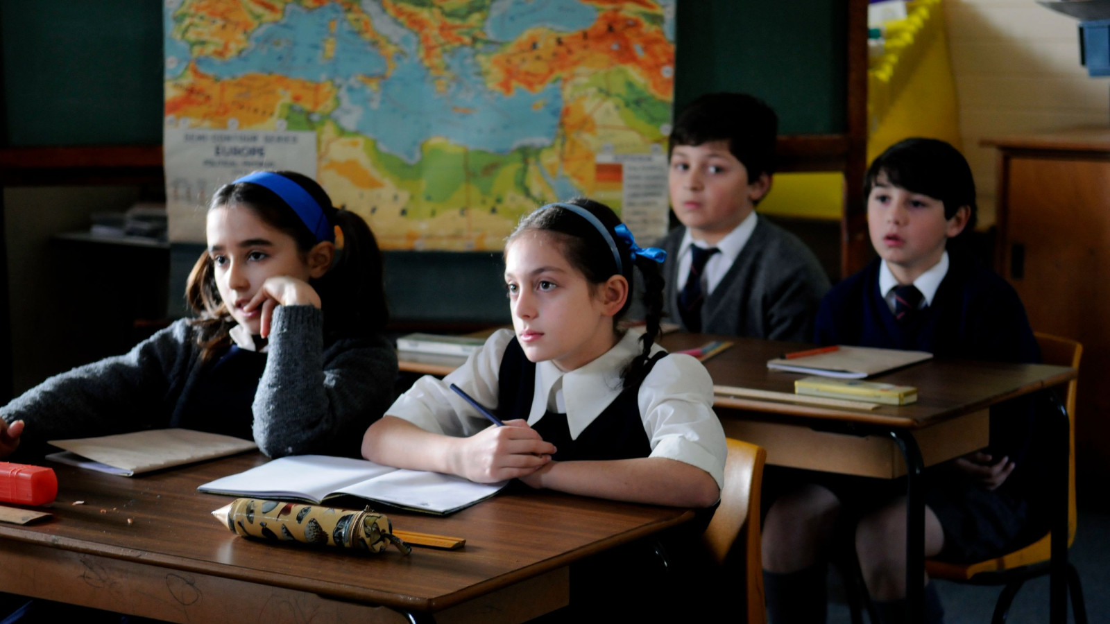A group of four school children sits at wooden desks in a classroom, looking attentively toward the front. A large world map is displayed on the wall behind them, and their desks are scattered with notebooks and stationery. The children wear school uniforms, with two girls having their hair tied back with blue headbands.