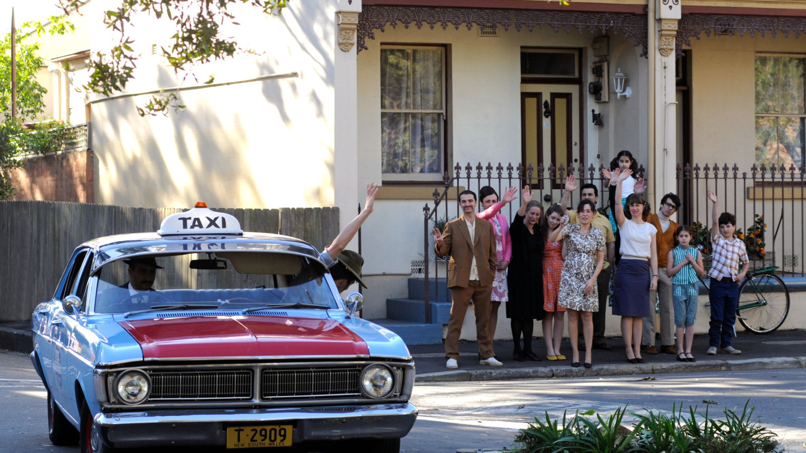 A blue and red taxi with a "TAXI" sign is parked on a street, while a diverse group of people, including men, women, and children, wave enthusiastically from the sidewalk in front of a two-story house. The scene captures a cheerful moment.