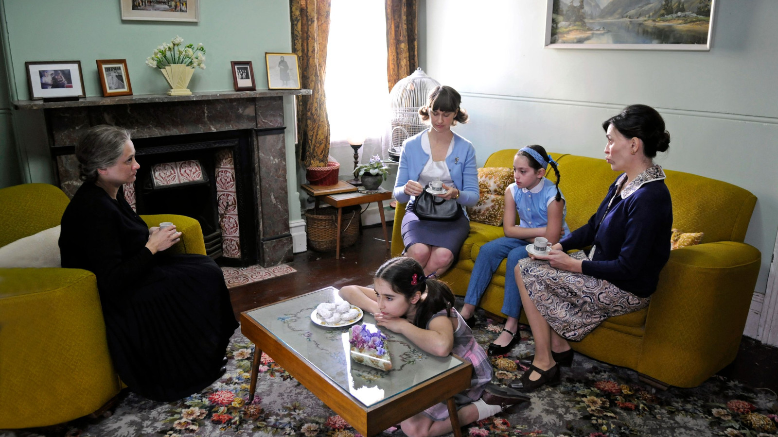 Five women of varying ages gather in a cozy living room, engaged in conversation over tea. The adults sit on a yellow couch while two young girls interact playfully on the floor, surrounded by patterned rugs and vintage decor, including a fireplace and photographs on the walls.