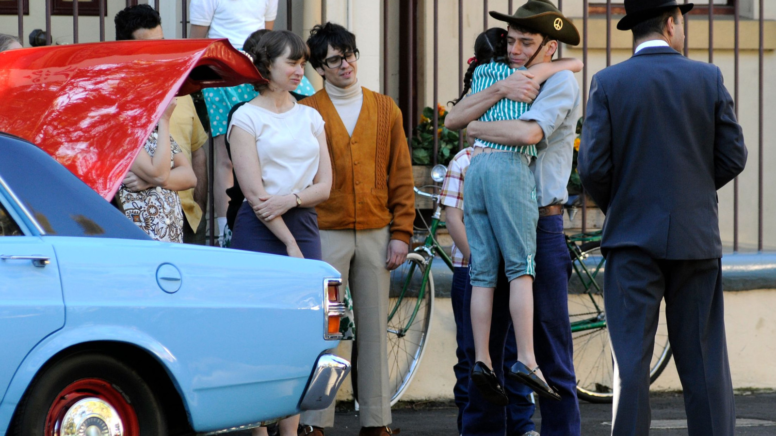 A group of people stands on a street, gathered around a blue car with its hood up. A man in a cowboy hat is holding a young girl, while others observe, including a woman with a relaxed expression and a boy wearing glasses. The background features a bicycle and a decorative fence.