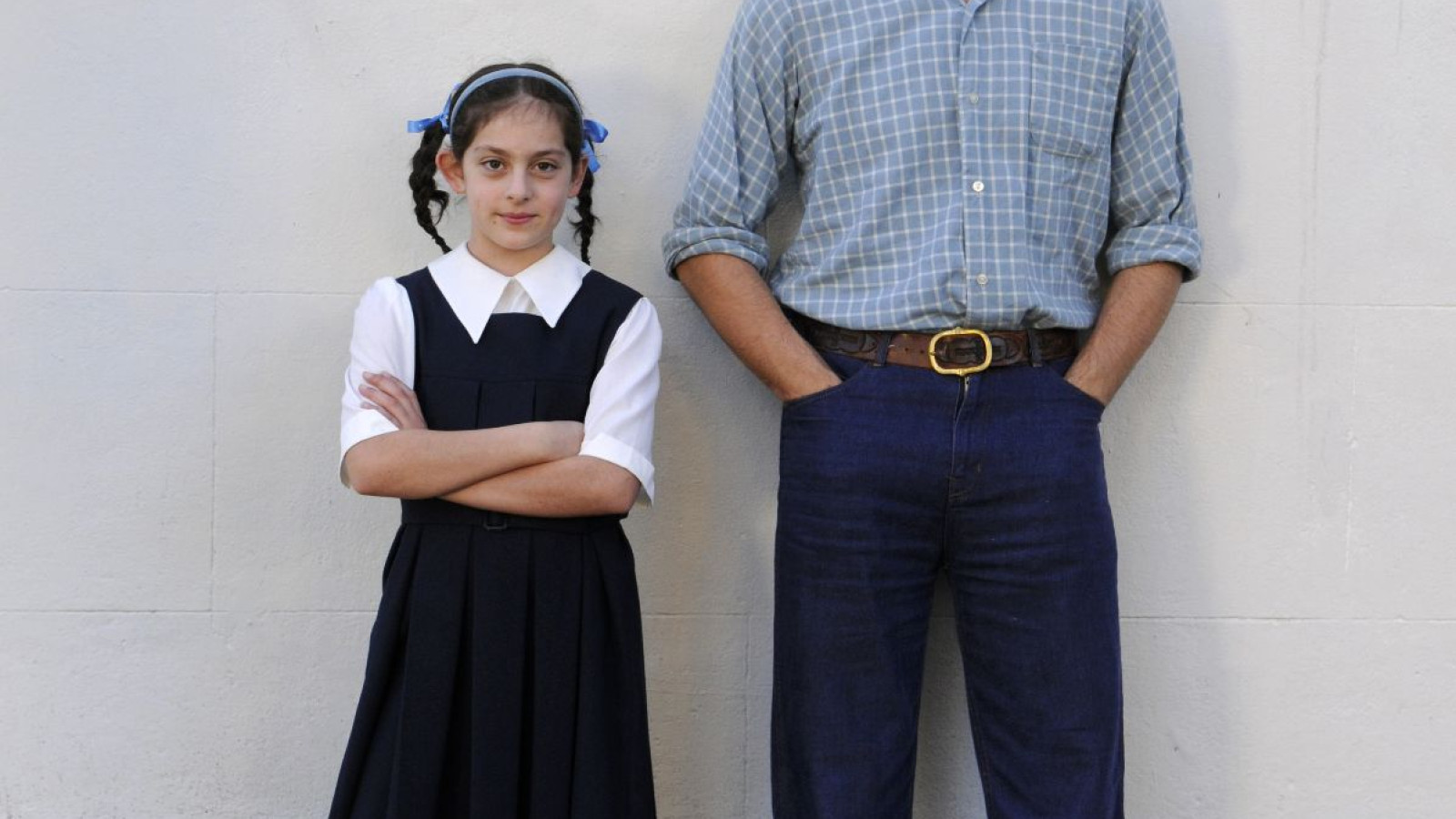 A young girl in a black school uniform with a white shirt and blue ribbons stands with her arms crossed, next to a young man in a checked shirt and dark jeans, smiling. They are positioned against a plain white background.