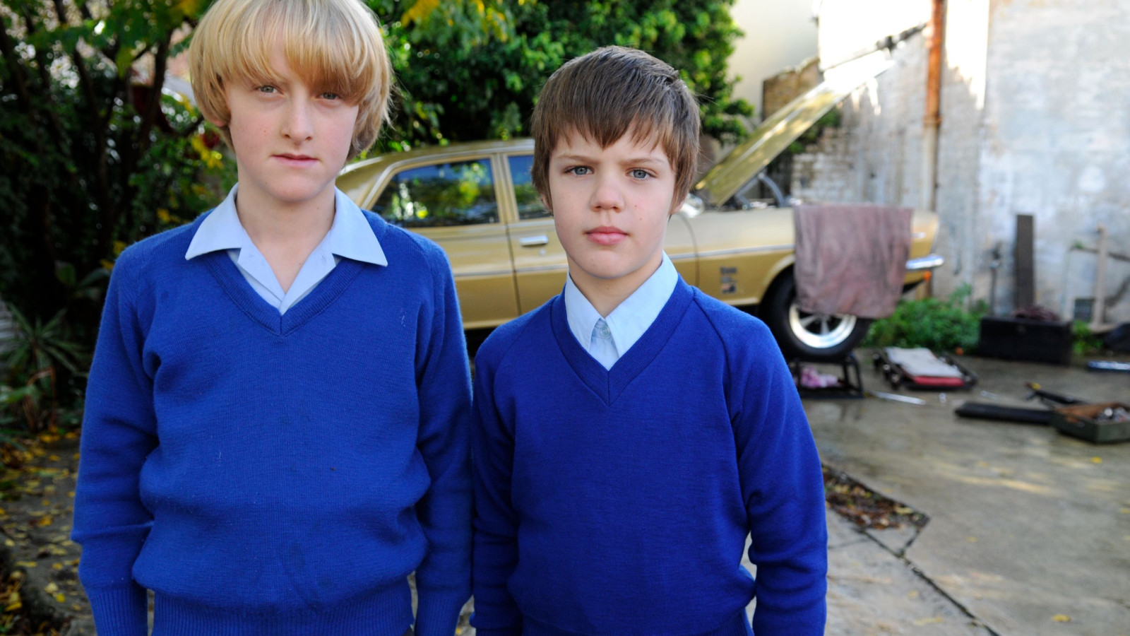 Two boys wearing blue sweaters stand together in an outdoor setting. Their serious expressions contrast with the vibrant greenery and a vintage car in the background, which has its hood raised. Various tools and parts are scattered on the ground nearby.