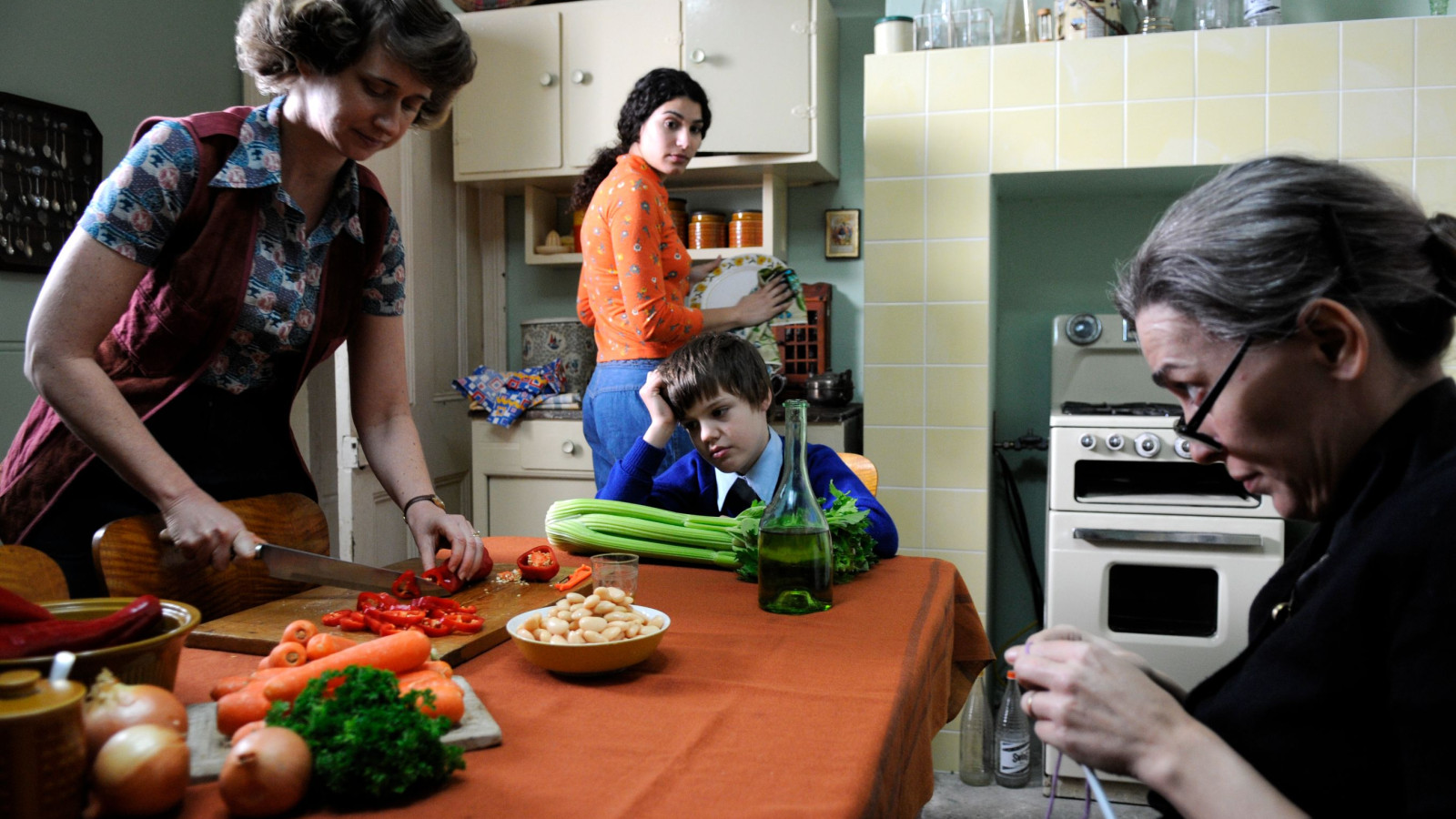 A woman cuts vegetables at a table, while another woman in an orange sweater stands nearby holding a plate. A boy in a blue sweater looks frustrated, resting his head on his hand, and an older woman in black is seated, knitting. Various ingredients are laid out on the table, including vegetables and a bottle of olive oil, with a retro kitchen background.