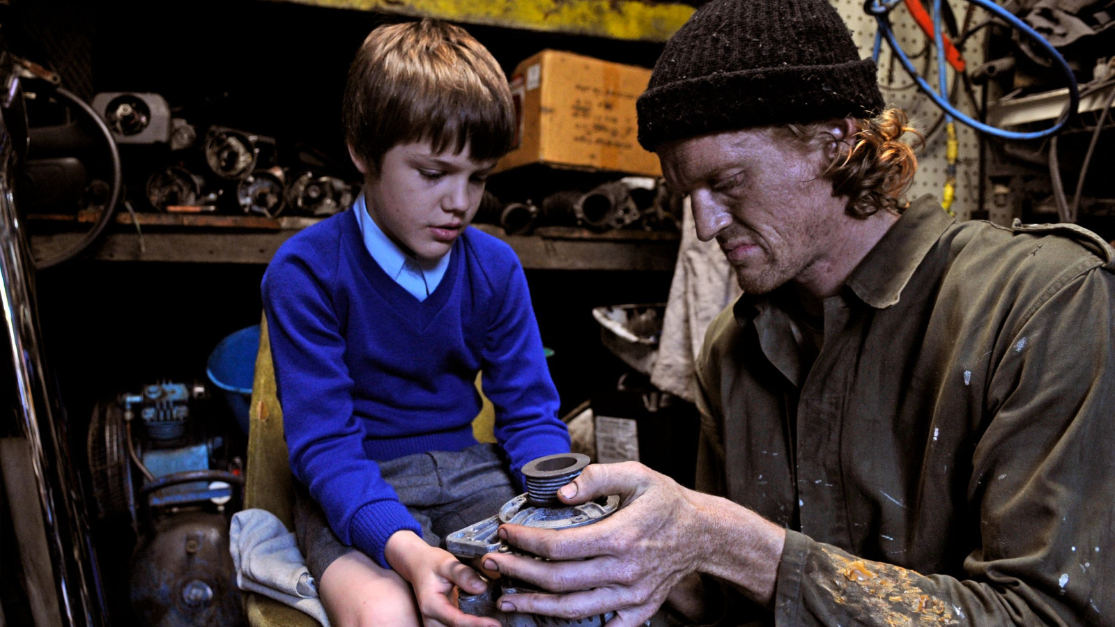 A young boy in a blue sweater and shorts sits on a chair, focused on a mechanical part held by a man dressed in a green jumpsuit and black beanie. The background is cluttered with various tools and parts, indicating a workshop environment. Both appear engaged in a learning moment, with the man sharing knowledge with the boy.