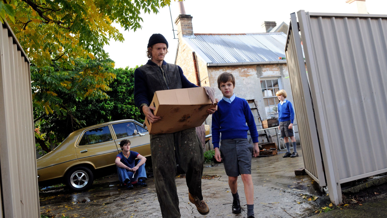 A man carrying a cardboard box walks through a gateway, accompanied by a boy in a blue sweater. In the background, another boy sits on the ground near a vintage car, while a third boy stands beside a building with a weathered facade. The scene is set outdoors, surrounded by greenery and scattered leaves.