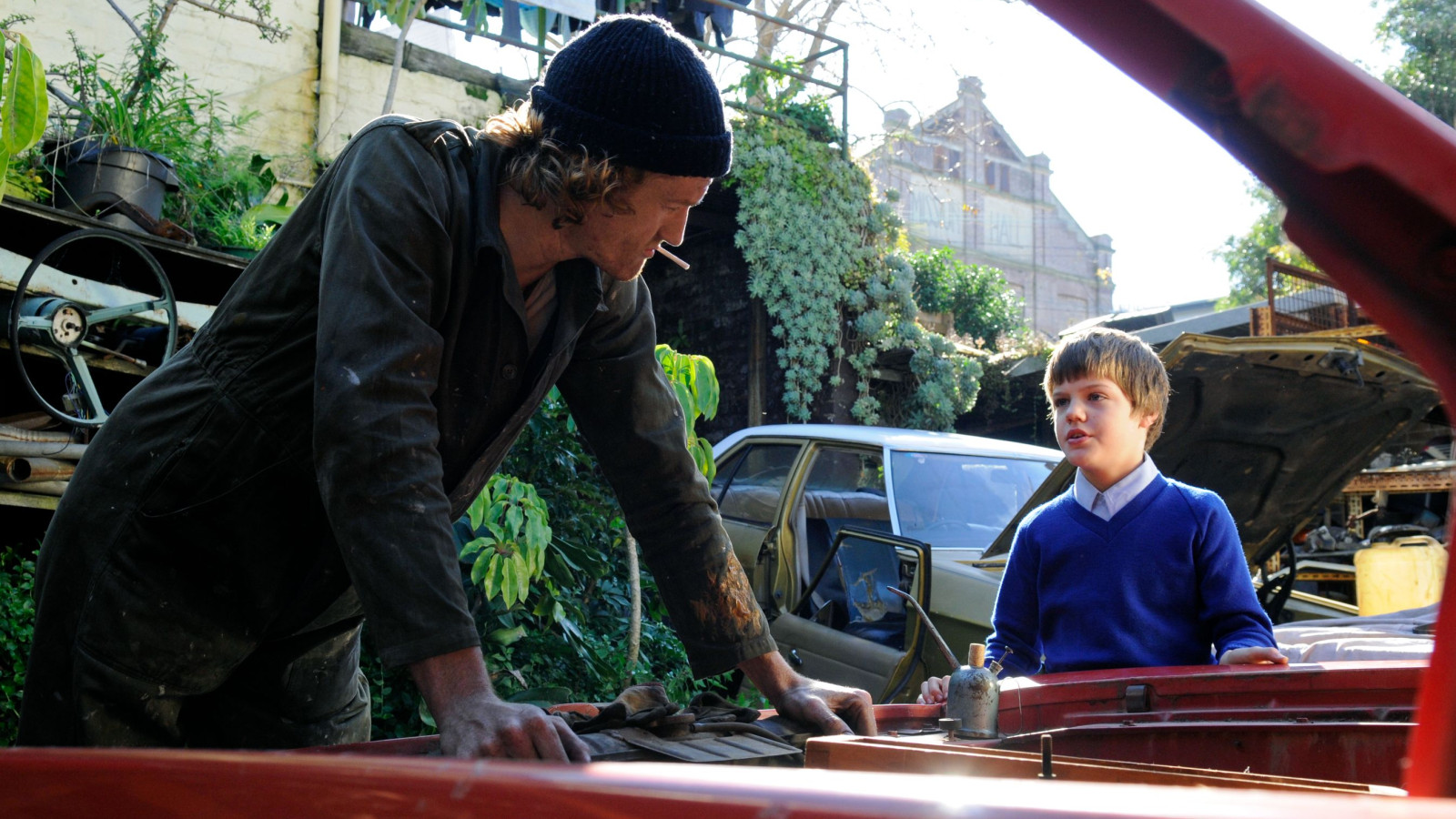 A man in a dark, dirty outfit leans over a car engine, speaking intently to a young boy wearing a blue sweater and white shirt. The background features overgrown plants and cluttered cars, creating an atmosphere of a makeshift workshop.