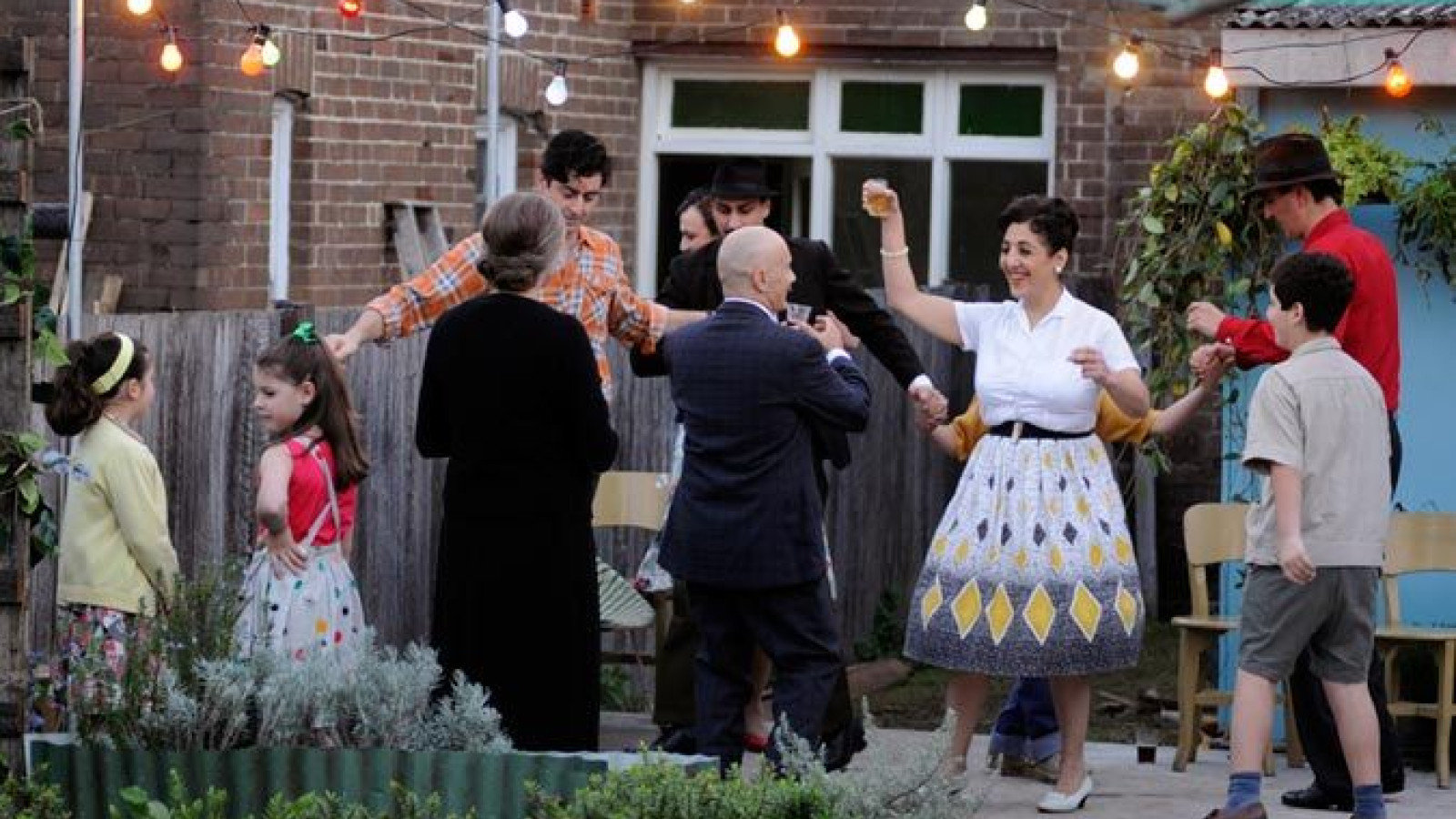 A lively outdoor gathering shows a group of people dancing and enjoying each other's company under string lights. The scene includes adults and children, with some dressed in vintage clothing, creating a cheerful and nostalgic atmosphere.