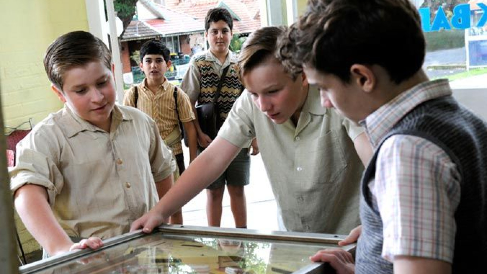 Three boys are gathered around a pinball machine, intently focused on the game. A fourth boy stands in the background, watching them with a curious expression. The setting appears to be a vintage arcade or game room, with visible greenery outside.