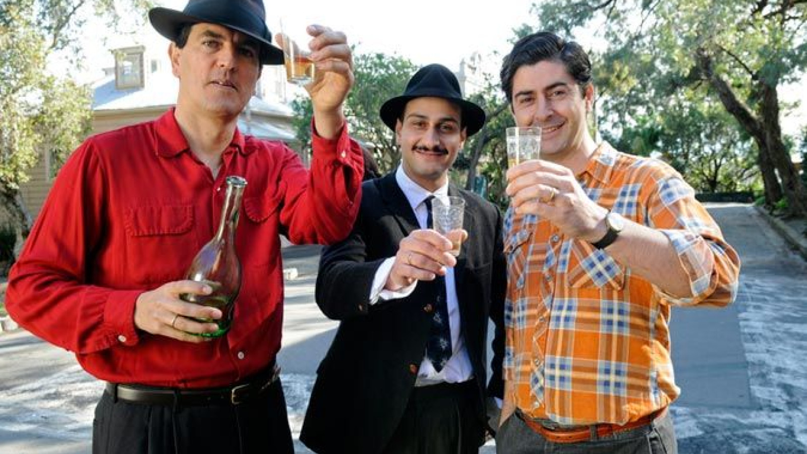 Three men stand outdoors, each holding drinks. One in a red shirt and black hat holds a bottle, the second in a black suit with a tie has a glass, and the third in an orange plaid shirt also raises a glass. They appear to be celebrating and smiling, with trees and buildings in the background.