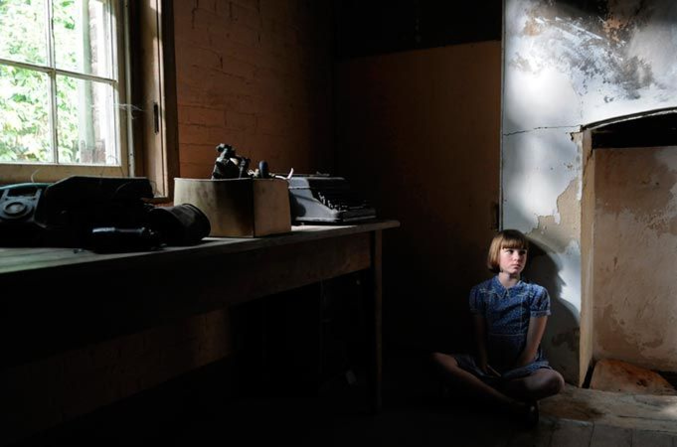 A young girl sits on the floor in a dimly lit room, leaning against a wall with a distressed surface. A wooden table nearby holds old typewriters and a box, while sunlight streams through a window, creating a contrast between light and shadow.