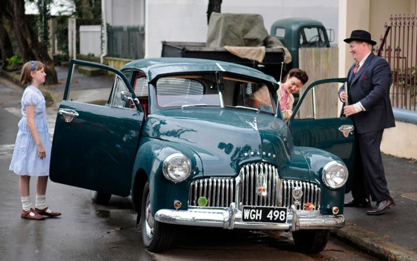 A vintage teal car is parked on a street, with its doors open. A young girl in a light blue dress stands next to the car, while a man in a suit and a hat, along with a woman in a floral dress, are engaging near the vehicle. The setting has a nostalgic charm, with trees and a house in the background.