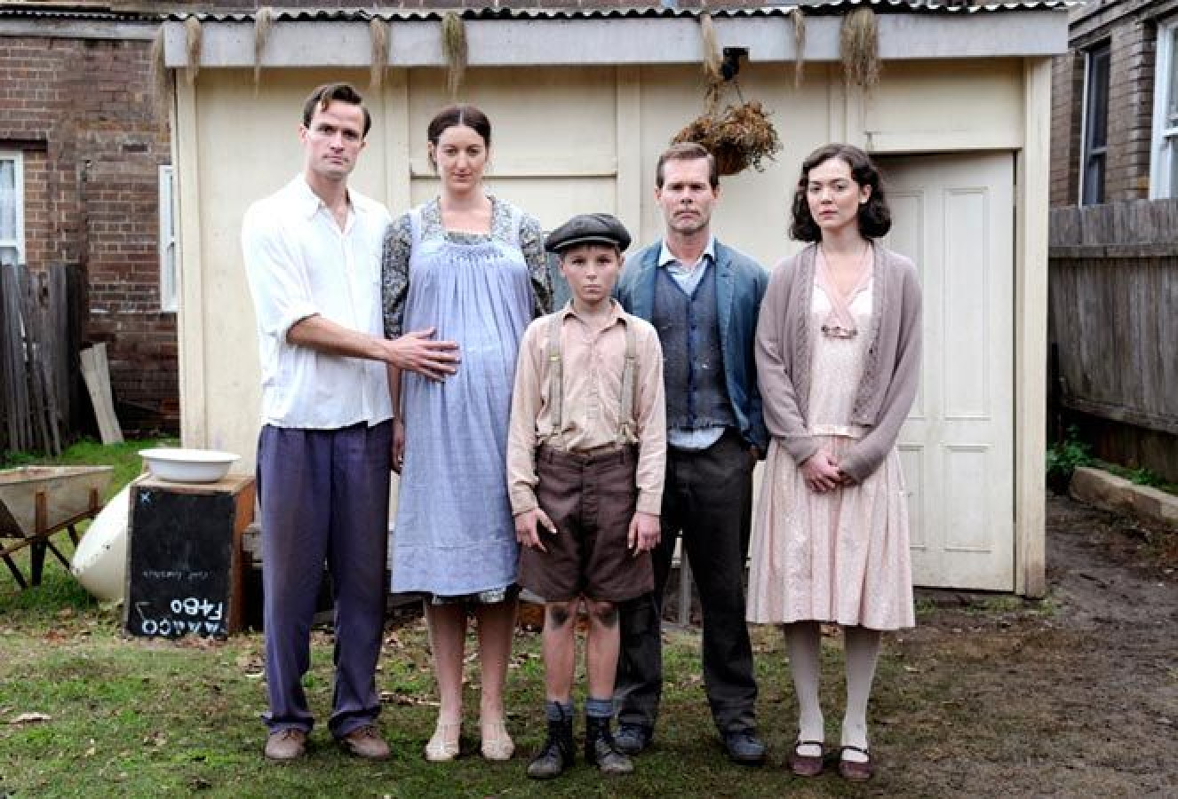A family stands outdoors in front of a small, weathered building. The group consists of five members: a man in a white shirt and blue trousers, a woman in a light blue dress, a young boy in shorts and a cap, a man in a grey vest, and a woman in a light pink dress. They all have serious expressions, with the man on the left resting his hand on the woman's shoulder. The background features a grassy area and a wooden fence.