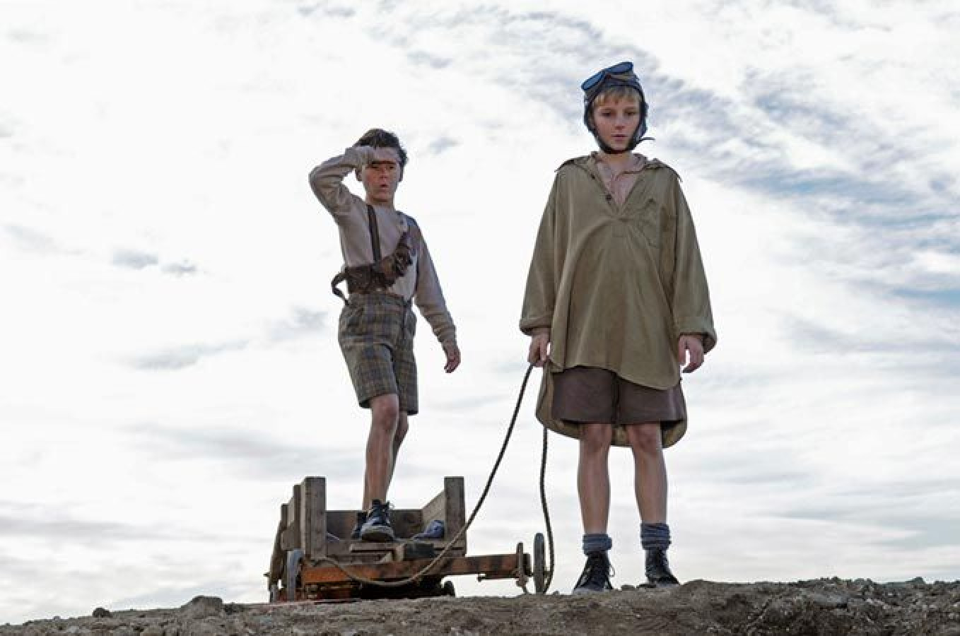 Two young boys stand on a hilltop against a cloudy sky. One boy salutes while the other, wearing a large coat and shorts, holds a rope attached to a makeshift wooden cart.