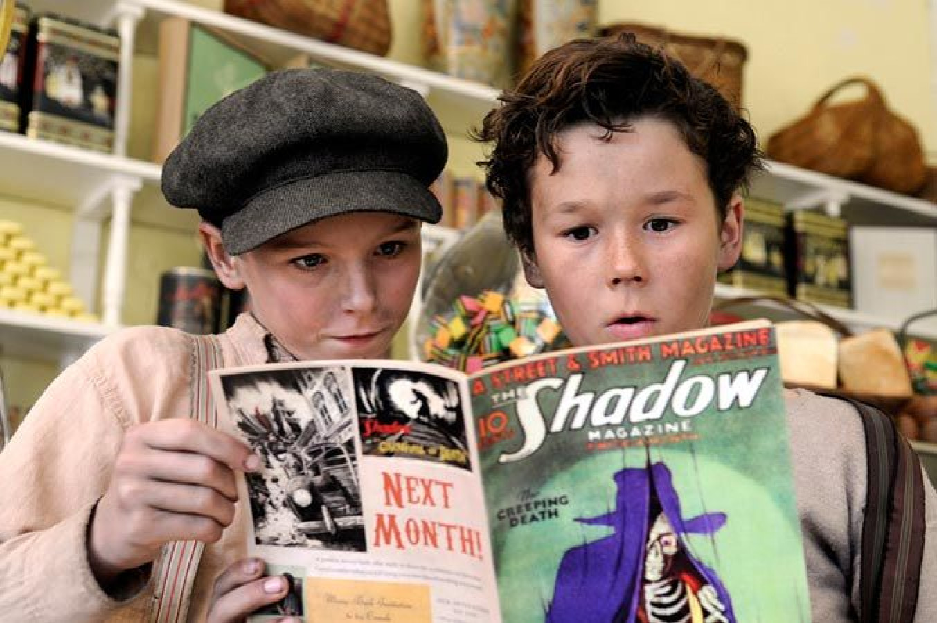 Two young boys closely examine an issue of "The Shadow" magazine. One boy wears a flat cap and a light-colored shirt, while the other has tousled hair and a serious expression, both appearing intrigued by the magazine's colorful cover. The background shows shelves filled with goods and candies.