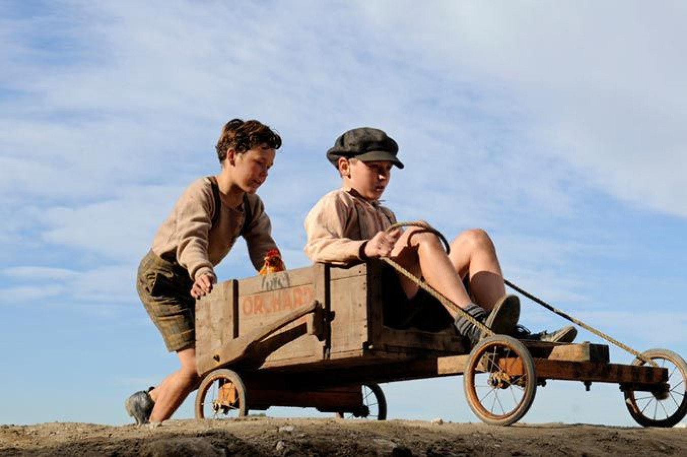 Two boys are outdoors, with one sitting in a wooden cart and holding onto a rope, while the other is pulling the cart along a dirt path. Both are dressed in simple, old-fashioned clothing, with a clear blue sky in the background.