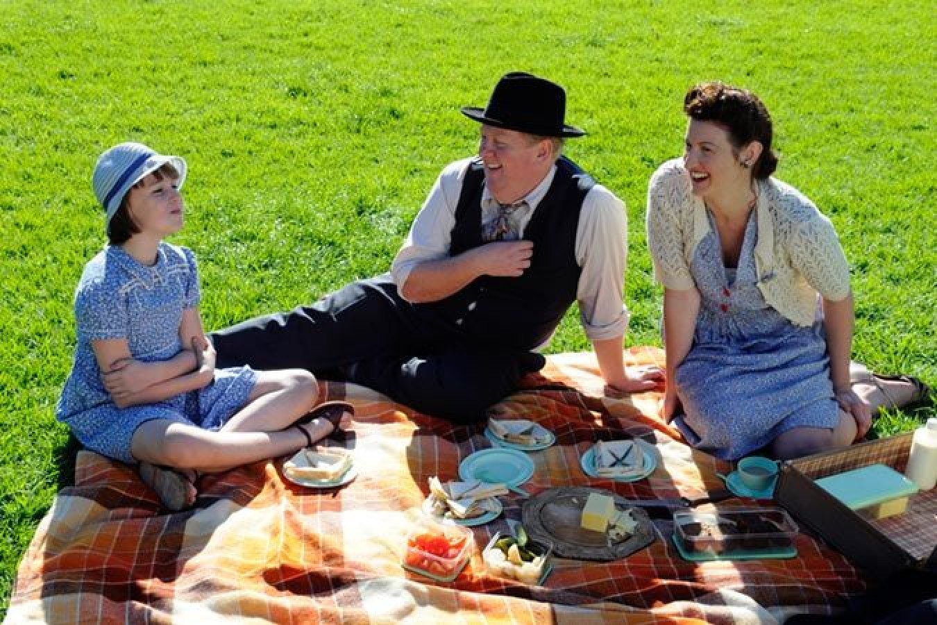 Three people enjoy a picnic on a blanket in a grassy park. A young girl in a blue dress and hat sits beside a man in formal attire, while a woman in a light sweater and dress laughs nearby. Plates of food are scattered around them.