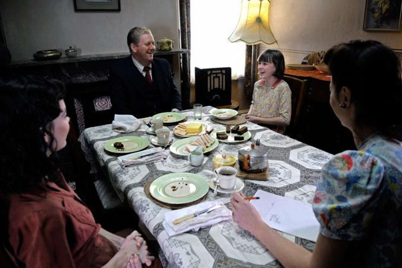 A dining table is set with plates of food, cups, and a teapot, surrounded by four individuals smiling and engaged in conversation. A young girl enjoys the moment, sitting across from a man in a suit, while two women, one in a blouse and the other in a dress, listen attentively. The room is warmly lit, with a lamp in the background and a cozy atmosphere.