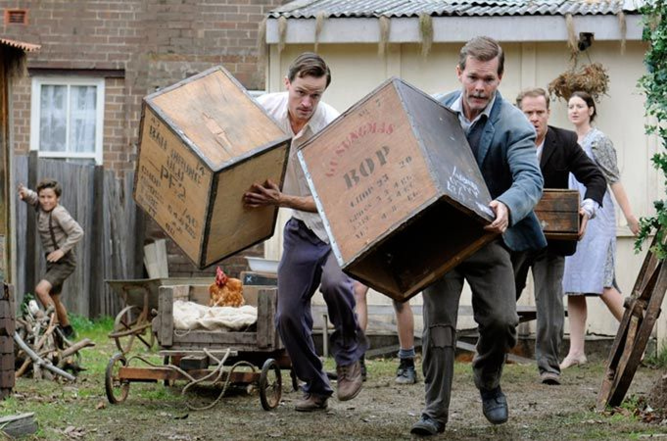 A group of people is moving large wooden boxes in an outdoor setting, with two men carrying heavy crates while a young boy watches from behind. Nearby, a woman stands holding another box, and a cart with a chicken is visible, alongside some scattered wooden items. The scene conveys a sense of urgency and teamwork.