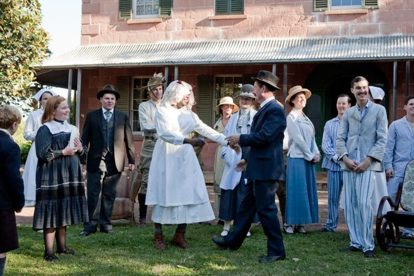 A group of people dressed in historical clothing gathers outdoors in front of a rustic, stone building. In the center, a man and a woman exchange a handshake, while other attendees observe and smile. The scene captures a lively, celebratory atmosphere, reminiscent of a bygone era.