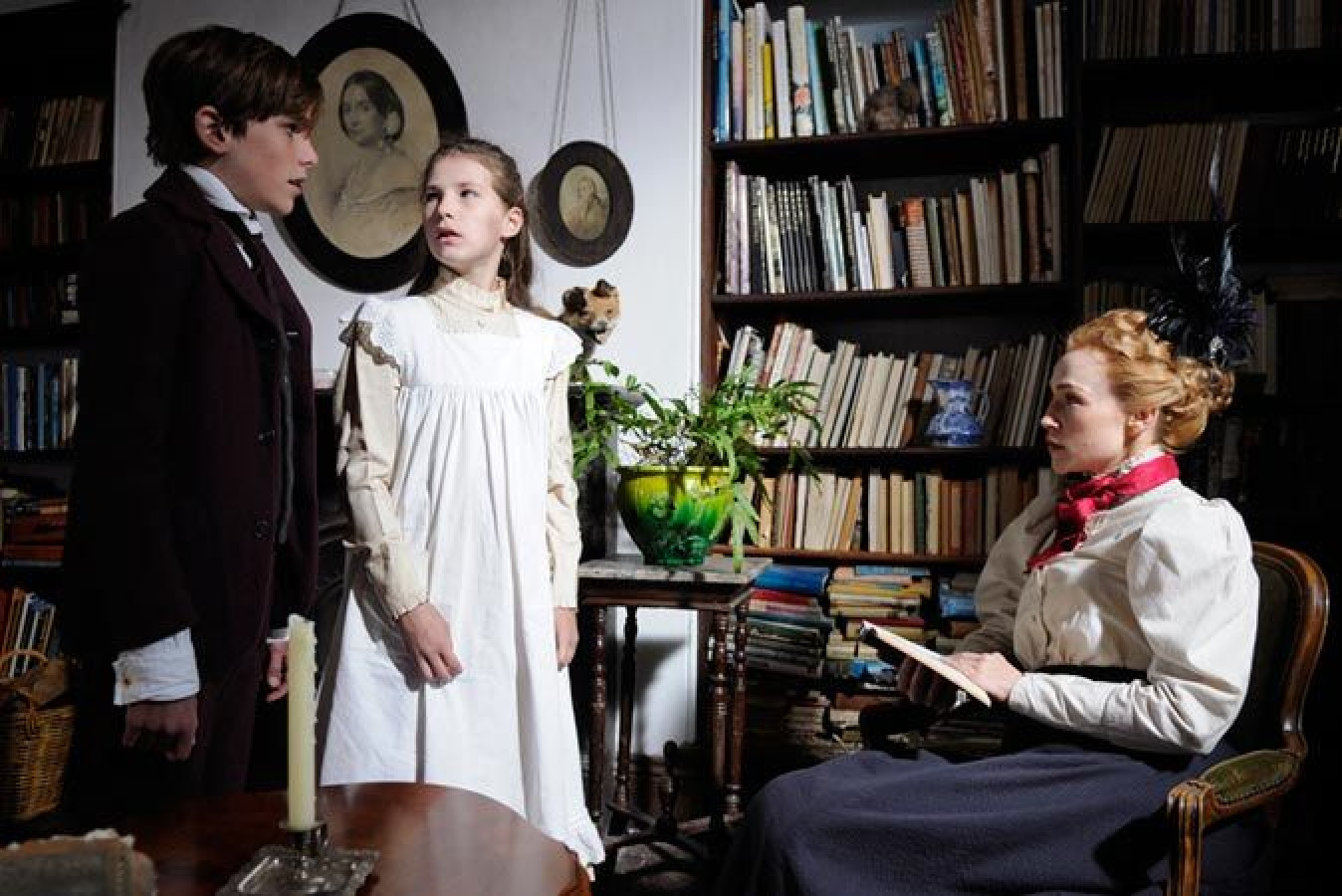A young boy in a dark suit stands beside a girl in a white dress, both appearing uncertain, as they look toward a woman sitting in a chair. The woman, dressed in period clothing, holds a book and gazes at them with an attentive expression. Behind them, a bookshelf filled with books and several framed portraits create a scholarly atmosphere. A green plant adds a touch of life to the scene.