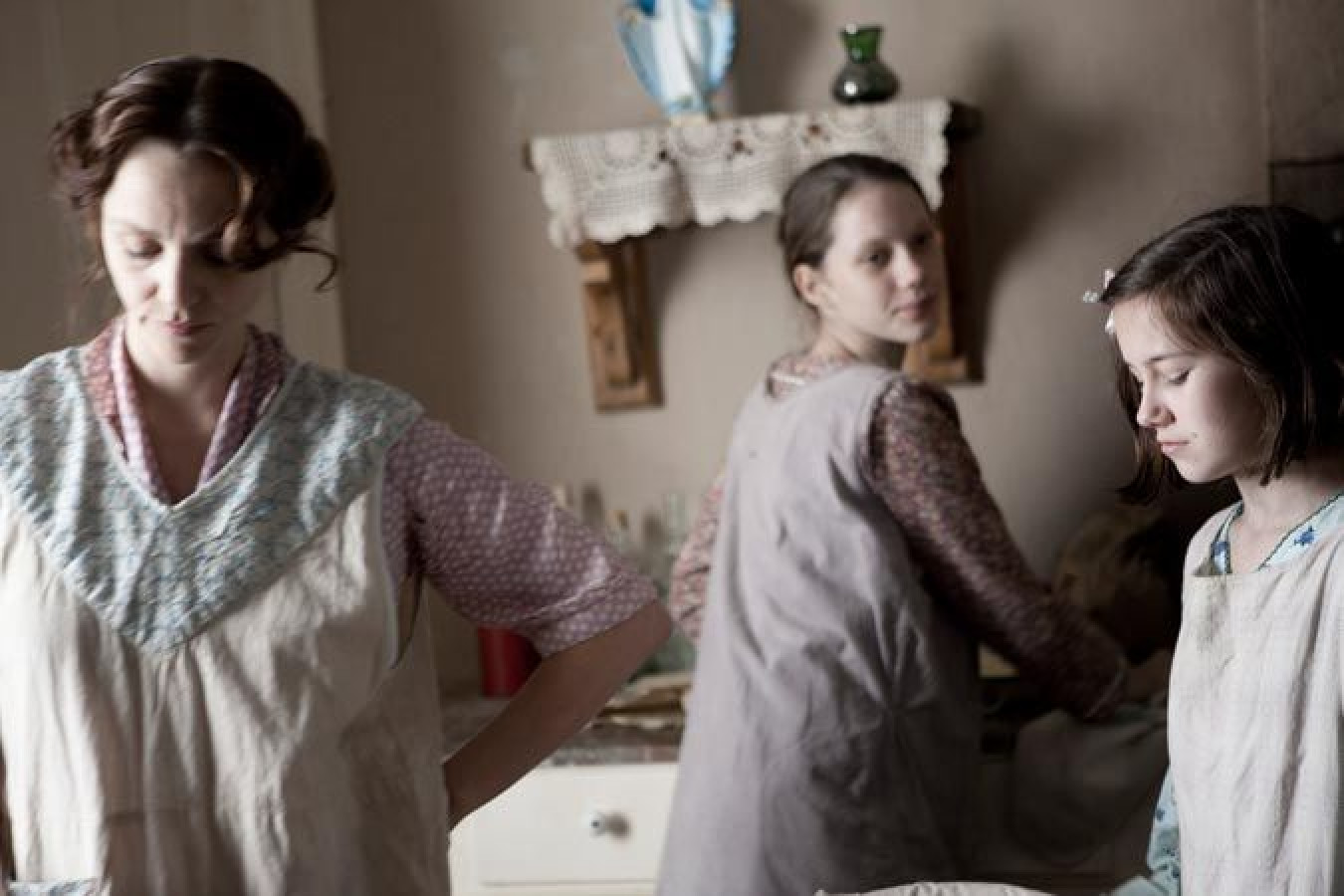 Three women are engaged in domestic tasks in a softly lit, rustic kitchen. The foreground features one woman in a blue and pink patterned top with a focused expression, while a teen girl in a patterned dress attends to a task nearby. Another young woman glances over her shoulder, creating a sense of camaraderie among them. The setting includes vintage kitchenware on shelves and a lace tablecloth, enhancing the atmosphere of a bygone era.