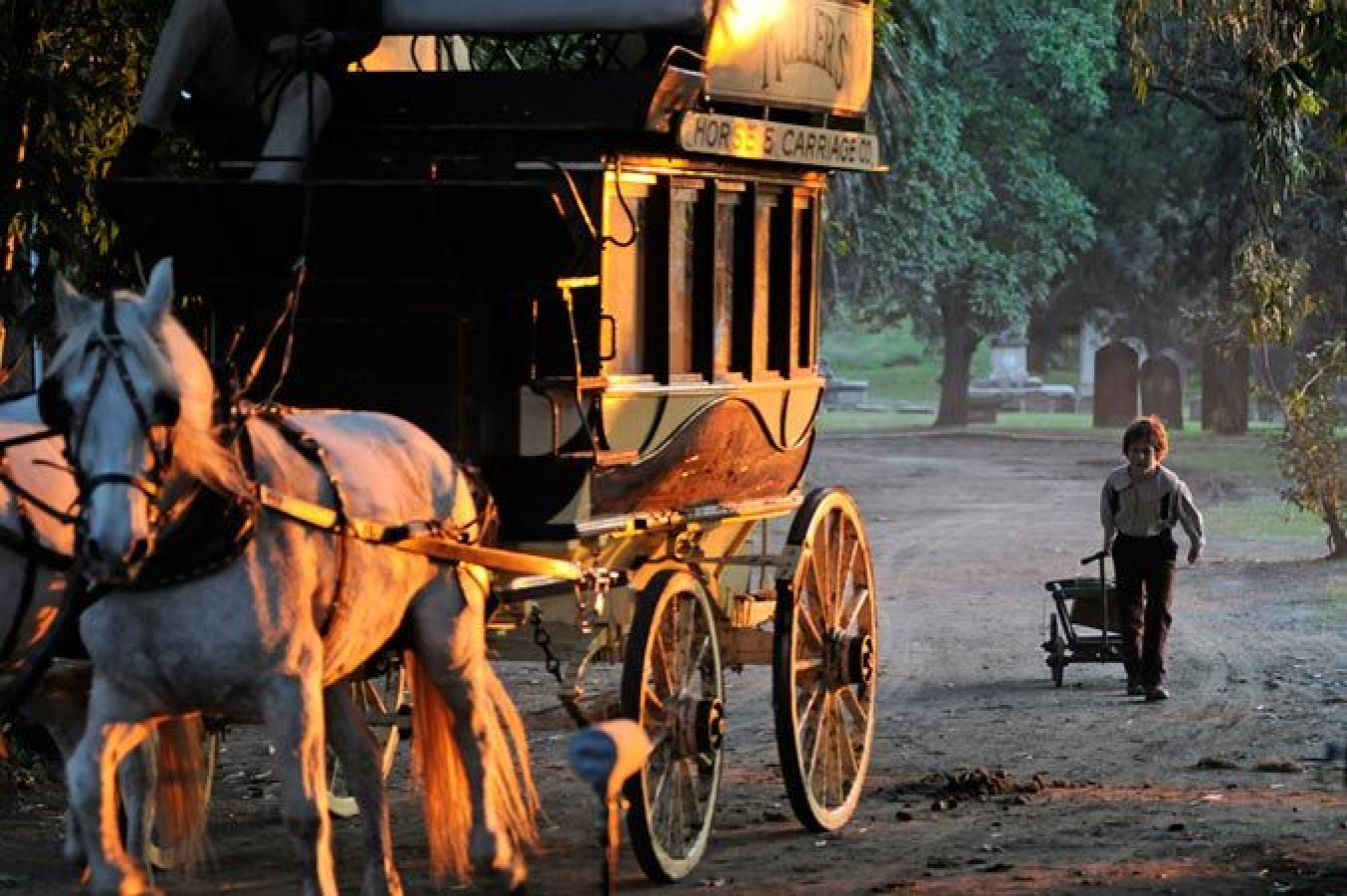 A horse-drawn carriage stands on a dirt path, illuminated by the soft glow of the sunset. A child walks beside the carriage, pushing a small cart, while trees and gravestones are visible in the background.