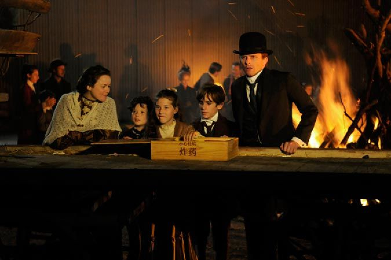 A group of five individuals stands near a large wooden table, illuminated by the glow of a fire in the background. Three children, a girl and two boys, are positioned in front of two adults, a woman in a shawl and a man wearing a formal suit and top hat. The scene conveys a historical setting with a gathering atmosphere.