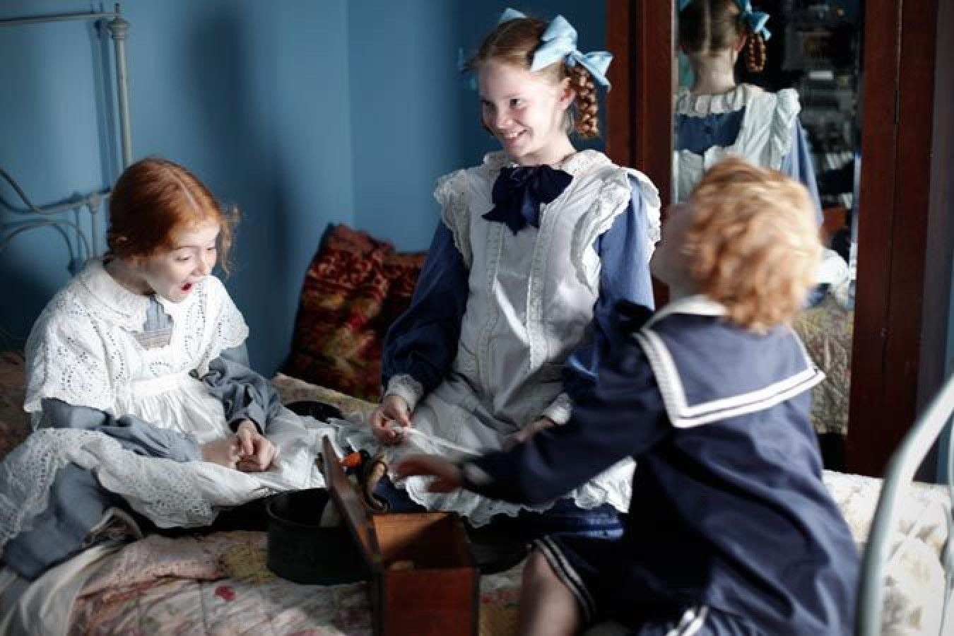 Three young children, dressed in vintage clothing, are sitting on a bed in a softly lit room. Two girls, one with auburn hair and the other with braided red hair tied with a blue bow, appear excited as they gather around a small wooden box. A boy with curly red hair, dressed in a sailor-style outfit, is reaching into the box, contributing to the joyous atmosphere. The room features a blue wall and a mirror reflecting their playful expressions.