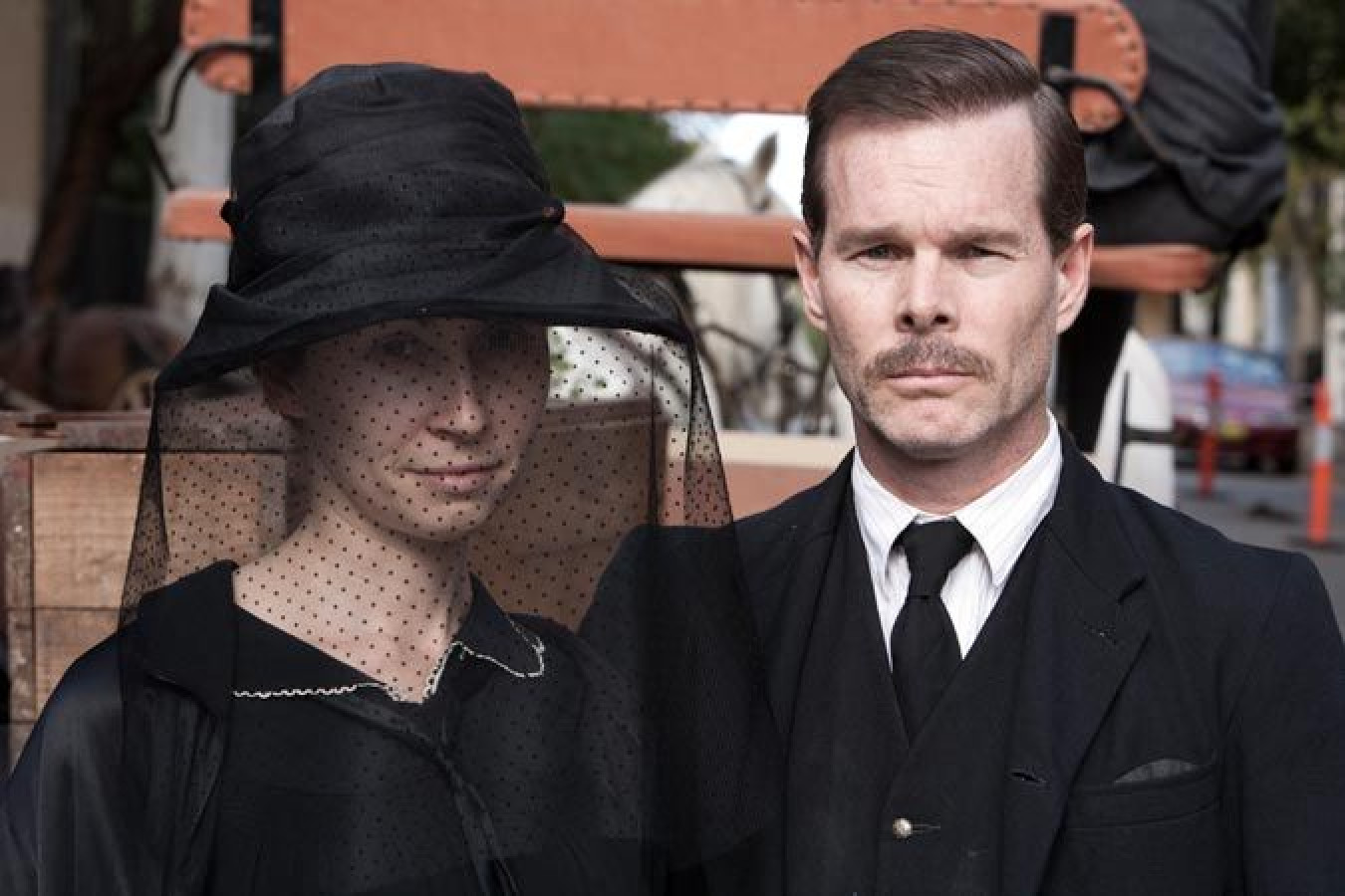 A man with a mustache wearing a classic black suit and tie stands beside a woman in a black dress and a wide-brimmed hat adorned with a sheer veil. They appear to be posing for a portrait, with a rustic background featuring a wooden cart and horses.