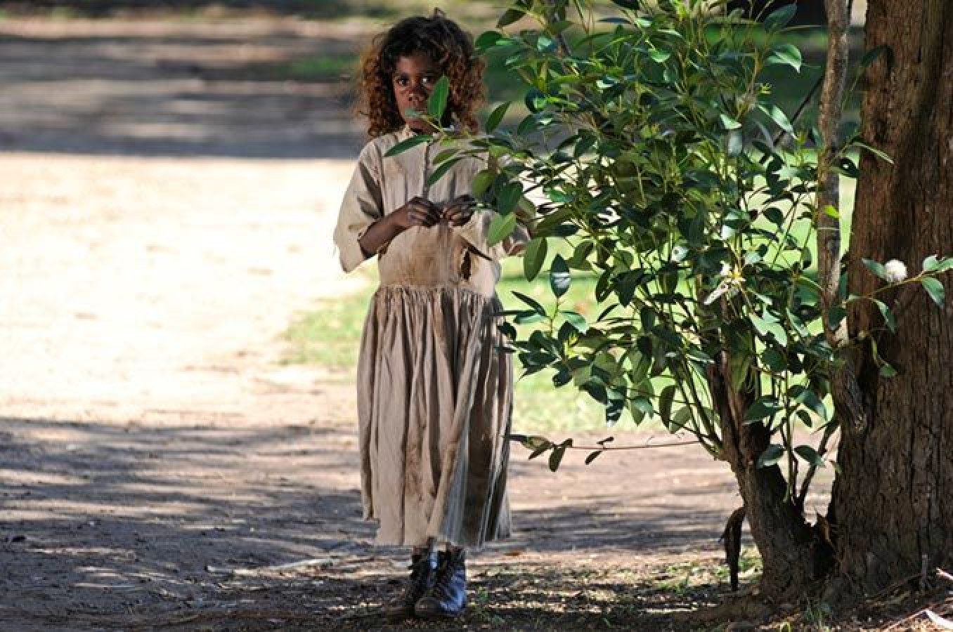 A young girl with curly hair, wearing a simple, faded beige dress, is standing near a bush, partially hidden. She appears to be holding something in her hands and looking intently at her surroundings.