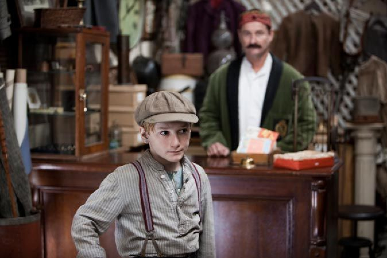 A young boy in vintage clothing and a cap stands in a shop, looking serious. In the background, a man with a mustache watches him, dressed in a green cardigan and white shirt. Various items and clothing are visible on shelves around them.