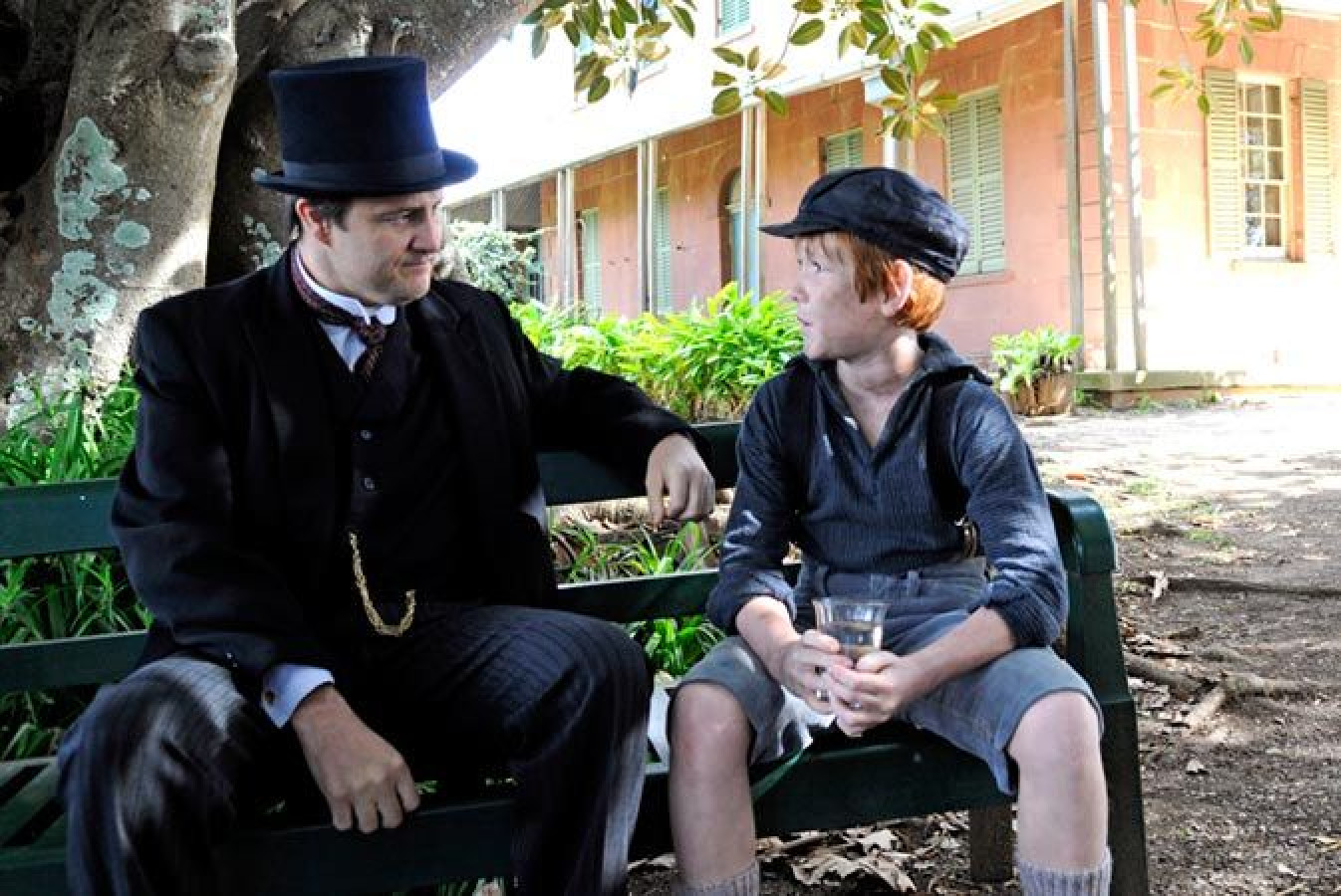 A man in a formal suit and top hat sits on a park bench, engaged in conversation with a boy dressed in period clothing, who holds a glass in his hands. Behind them, a lush green landscape and a building with shuttered windows create a historical setting.
