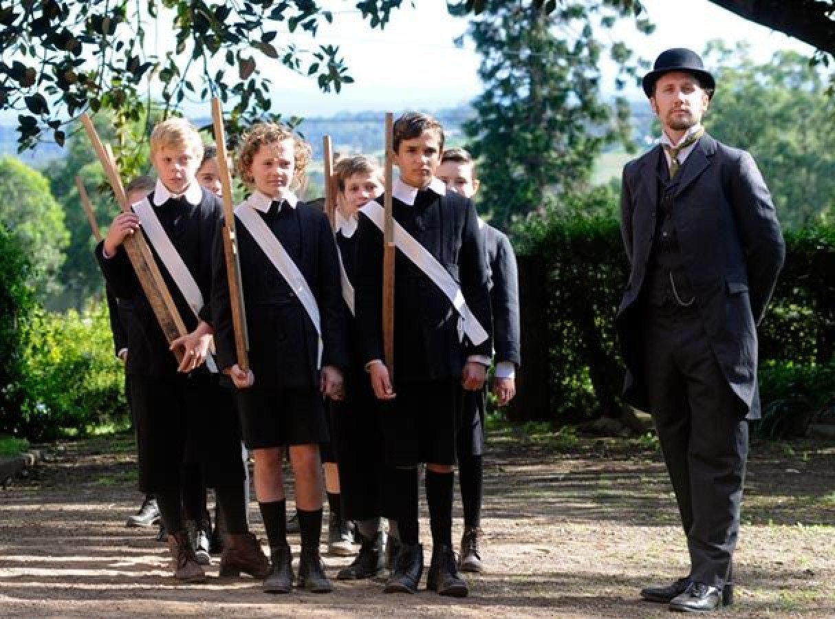 A group of boys dressed in early 20th century school uniforms stands in line, each holding wooden sticks. They wear white sashes across their torsos, while a man in formal clothing and a top hat stands slightly apart, observing them. The background features lush greenery and a bright, clear sky.