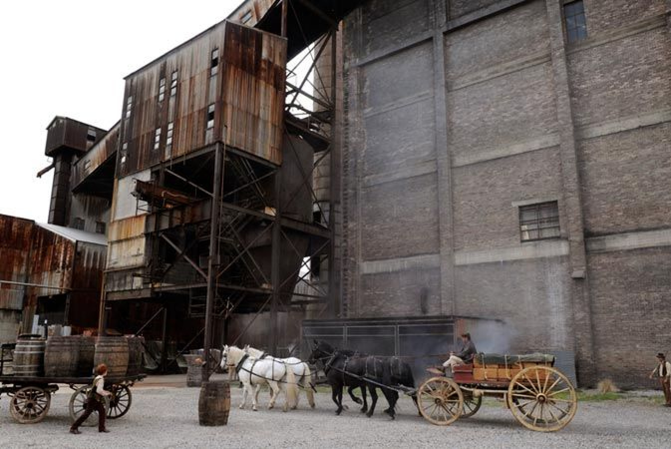 A weathered industrial building with rusted metal and brick exteriors looms in the background. In the foreground, a team of horses pulls a wooden cart while a person tends to barrels scattered around, surrounded by dust and smoke.