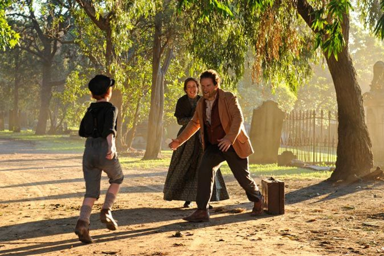A boy runs excitedly towards a man with a suitcase, while a woman smiles joyfully behind them in a sun-dappled park. The scene captures a warm reunion surrounded by trees and a slightly hazy background.